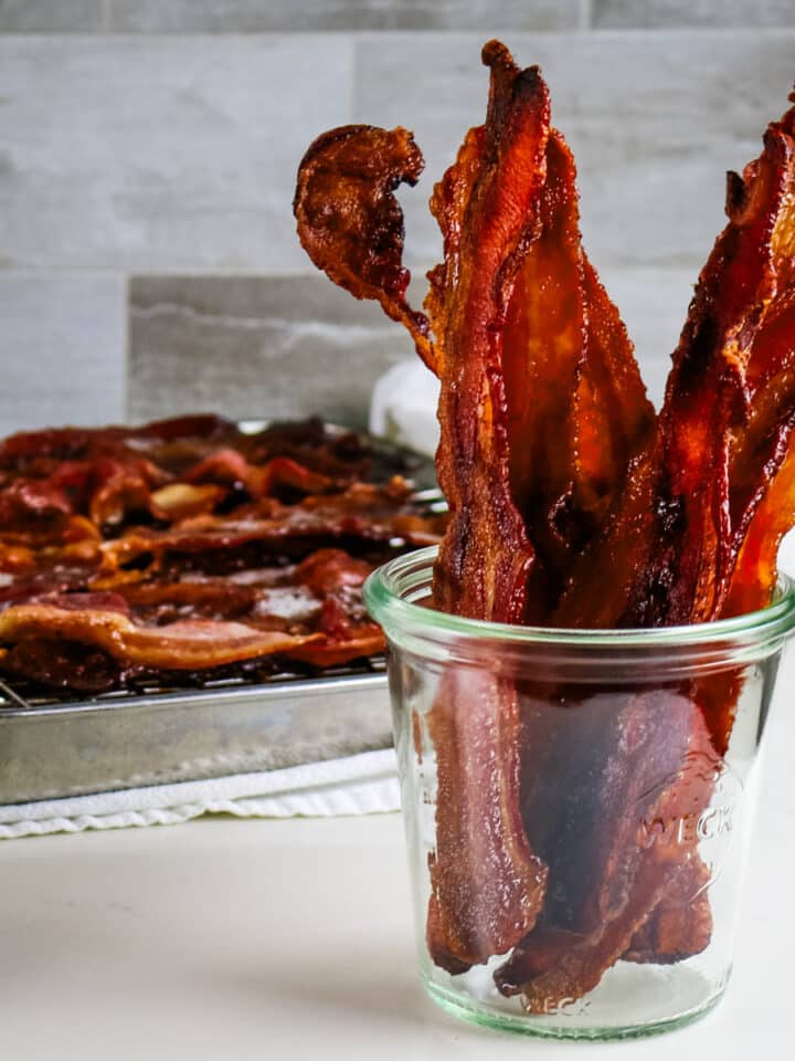 Crispy bacon strips in a clear glass jar, with more bacon cooling on a rack over a metal tray in the background.