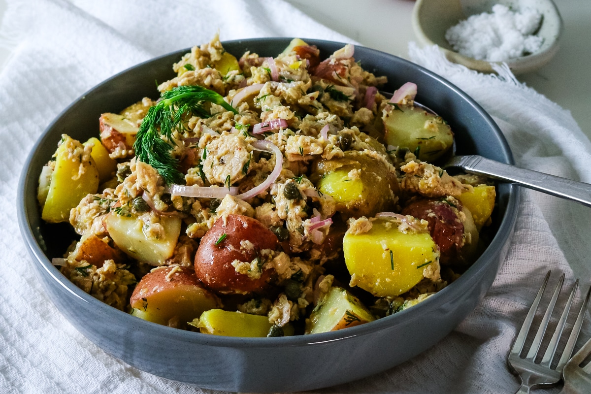 A gray bowl filled with tuna potato salad, with a spoon inside, and salt on the side.