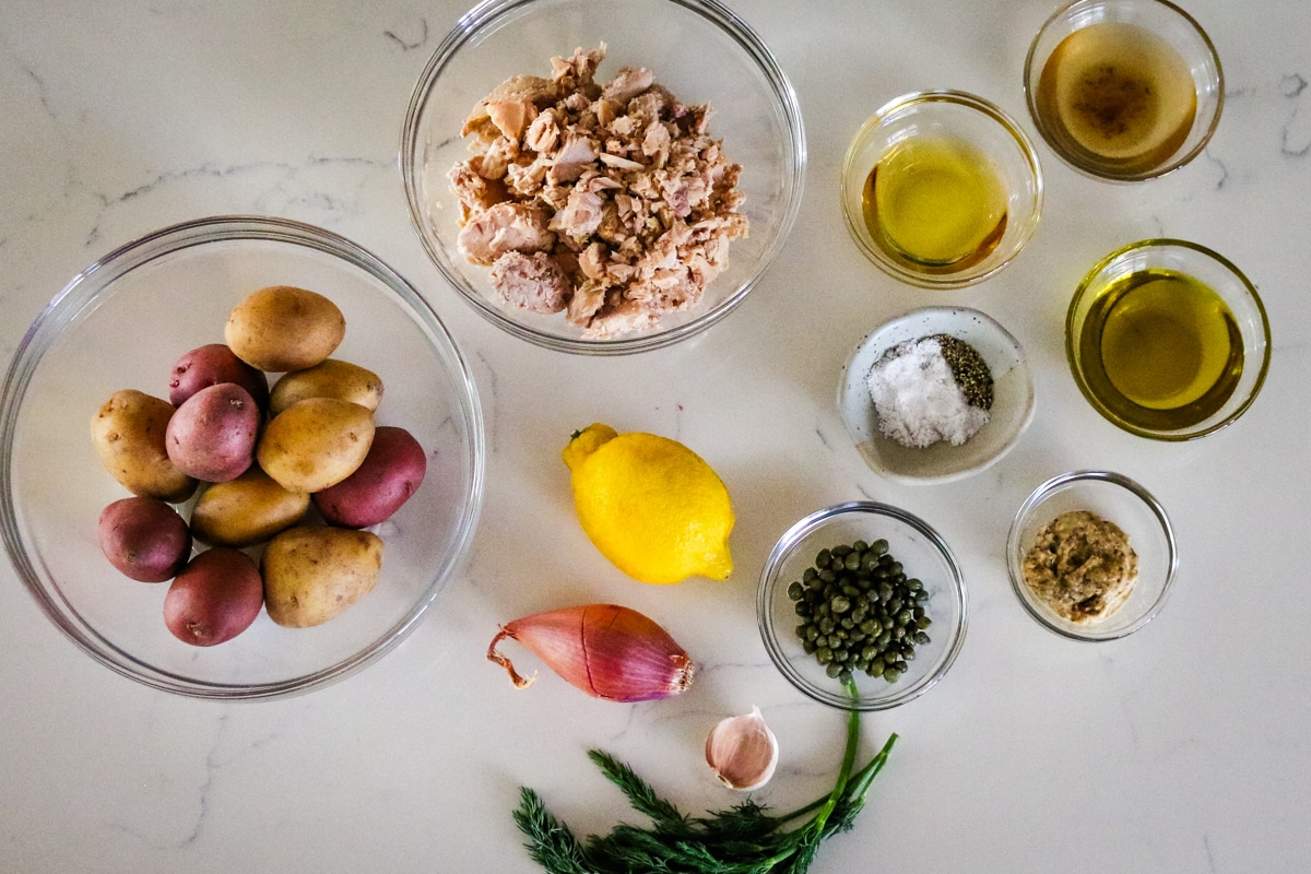 Potatoes, tuna, and seasonings in a glass bowl, with lemon, onion, a clove of garlic, and herbs on the table.
