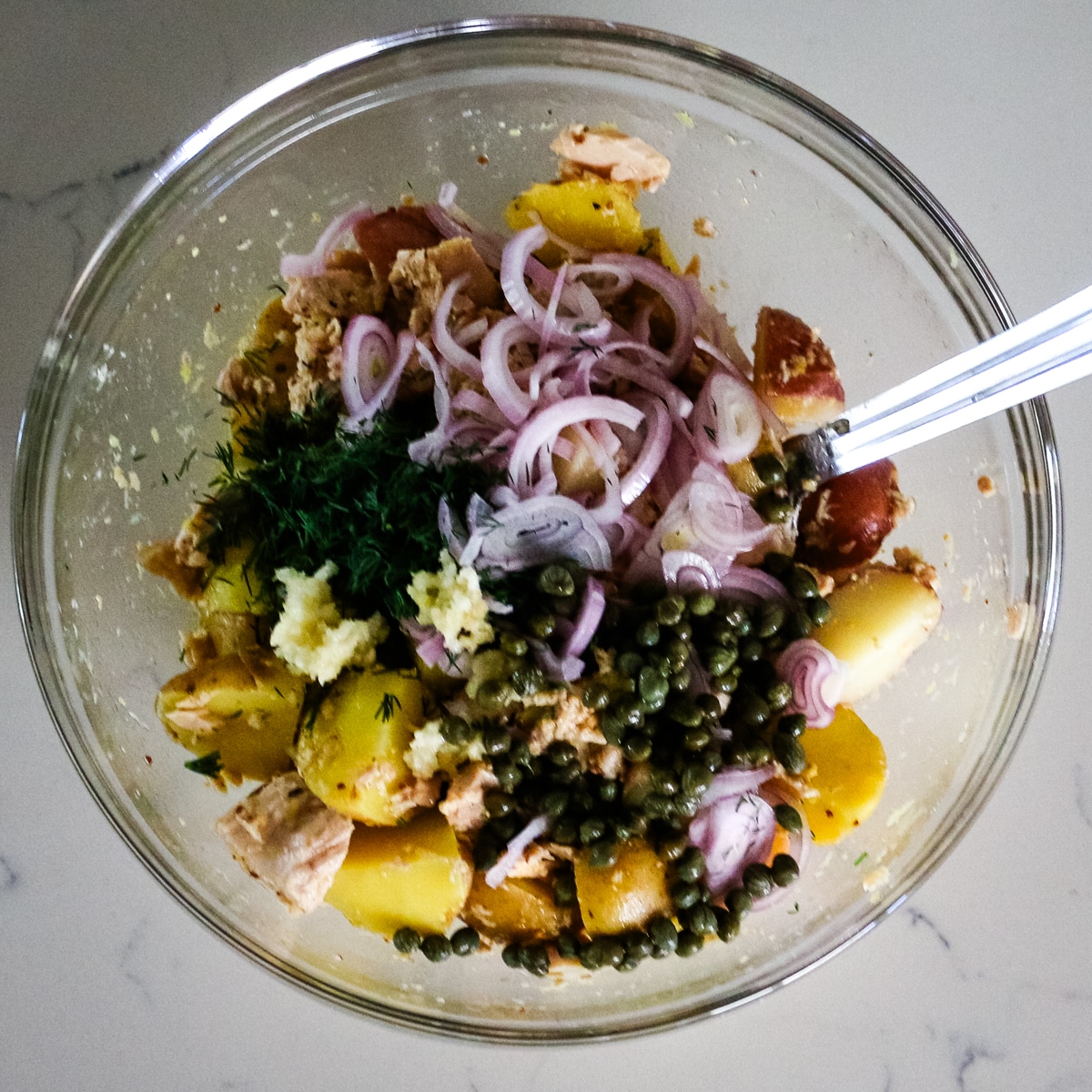 A clear bowl filled with a mix of tuna, potato, onion, herbs, and seasoning.