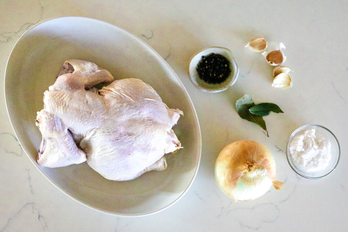 A whole chicken on a white plate, with black pepper, laurel leaves, a few garlic cloves, salt, and one onion on the table.