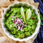 Chunky guacamole in a white bowl, with tortilla chips beside it on a white plate.