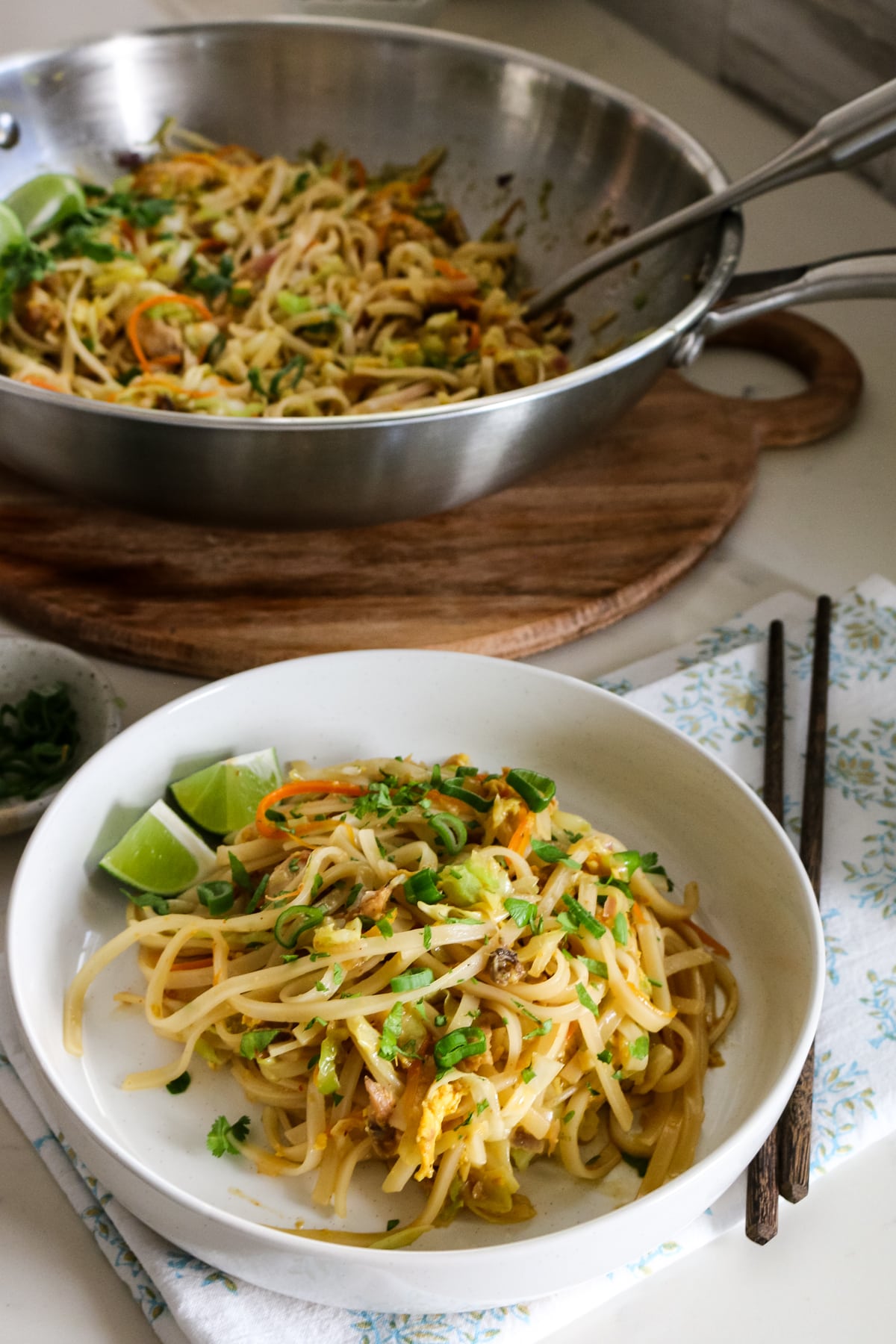 Chicken Pad Thai in a white bowl with chopsticks beside it, and a skillet of Pad Thai in the background.