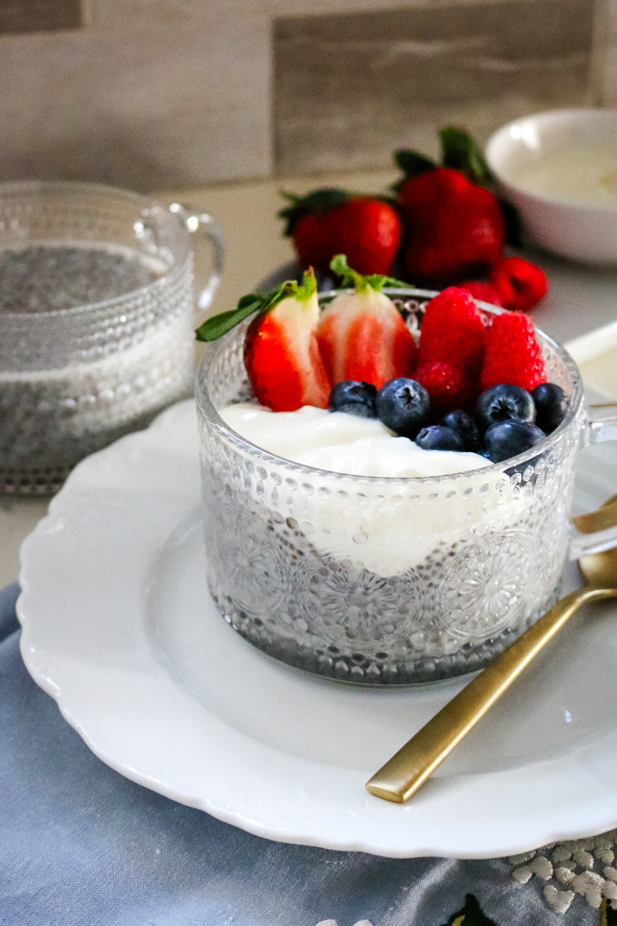 Two chia seed puddings in clear cups, one topped with berries, served on a white plate with a spoon.