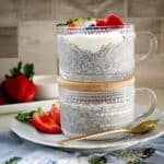 Chia seed puddings in clear cups, served on a white plate with a strawberry and spoon on the side.