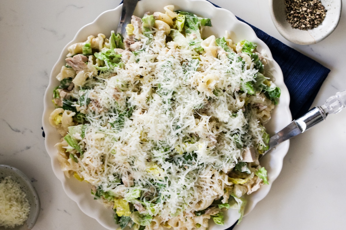 Chicken Caesar pasta salad on a white plate with a fork in it, served with black pepper and cheese on the side.