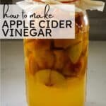 large glass jar filled with chopped apples and liquid for fermentation, covered with paper and secured with a rubber band.