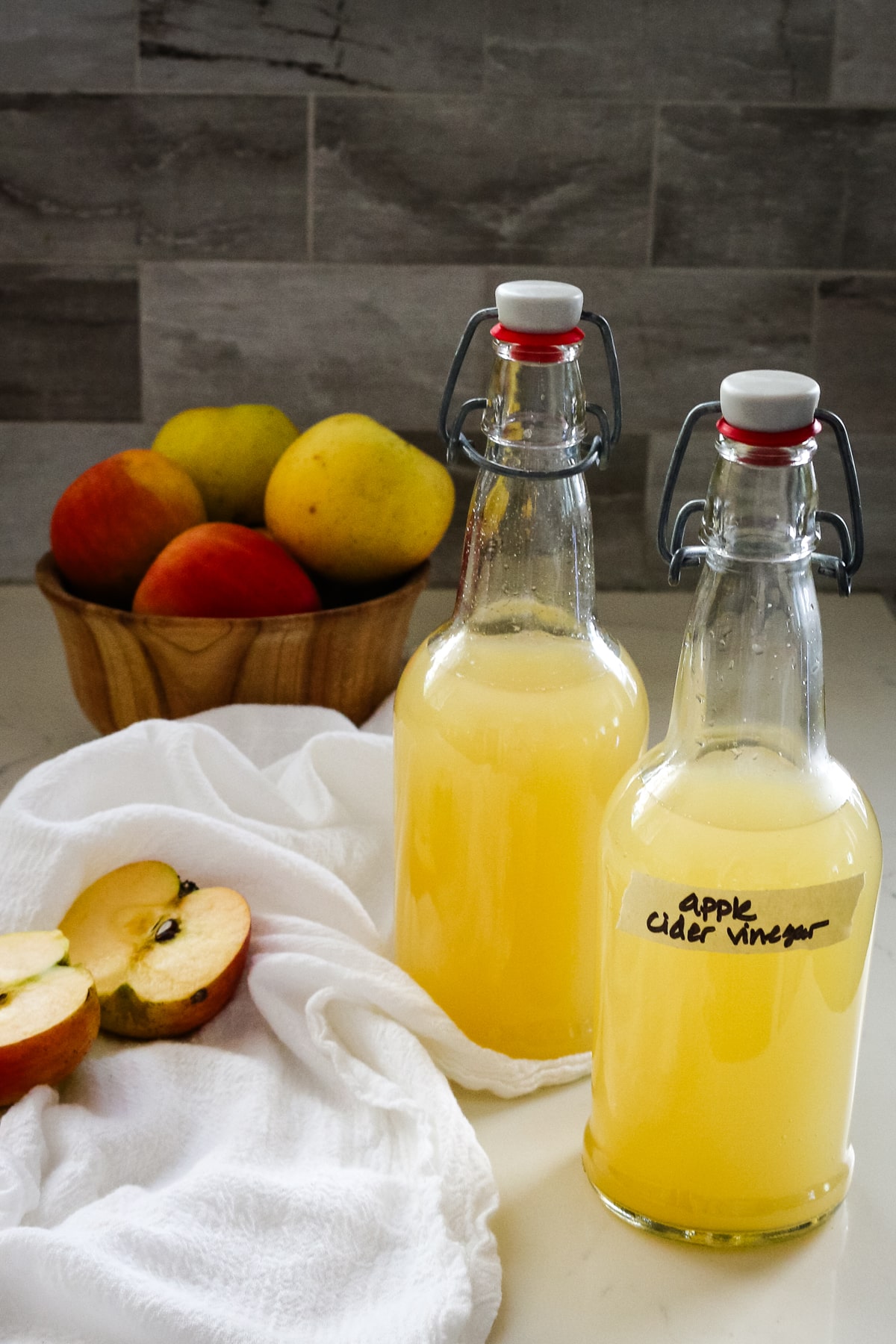 two swing-top bottles of homemade apple cider vinegar next to a bowl of fresh apples.