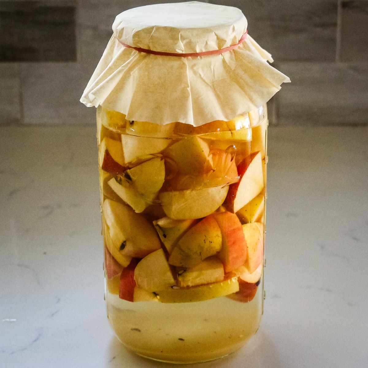 large glass jar filled with chopped apples and liquid for fermentation, covered with paper and secured with a rubber band.