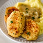 three golden-brown chicken kotleti served on a white plate with a side of mashed potatoes topped with gravy.
