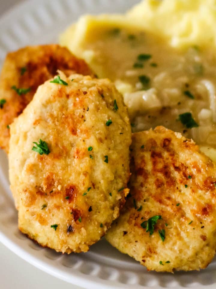 three golden-brown chicken kotleti served on a white plate with a side of mashed potatoes topped with gravy.