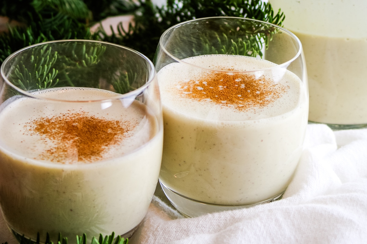 Two glasses of creamy eggnog sit on a white cloth, each topped with a sprinkle of cinnamon.