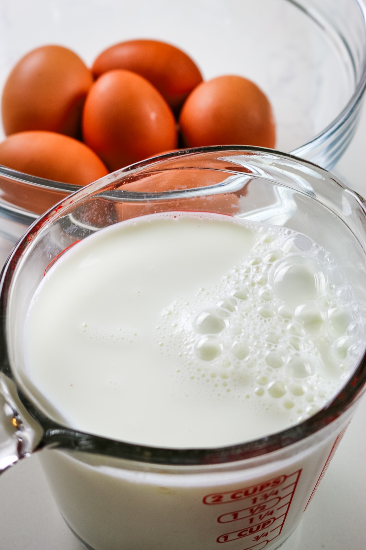 A glass measuring cup filled with milk sits in the foreground, with small bubbles on the surface. Behind it, a clear bowl holds several brown eggs.