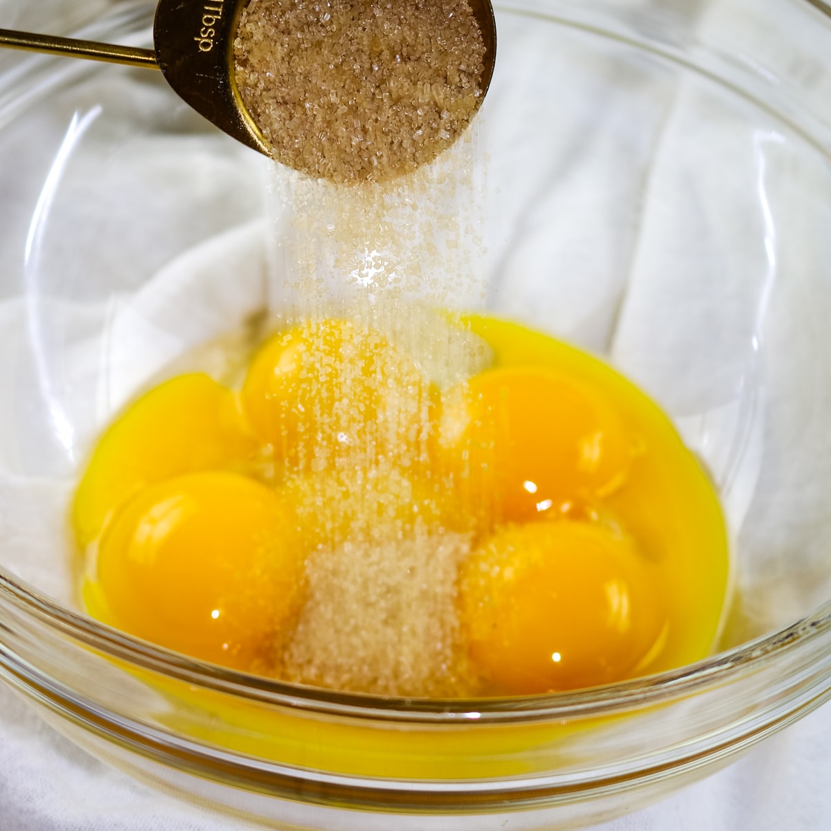 A close-up of a glass mixing bowl containing four raw egg yolks, with brown sugar being poured from a gold measuring spoon into the center of the yolks.