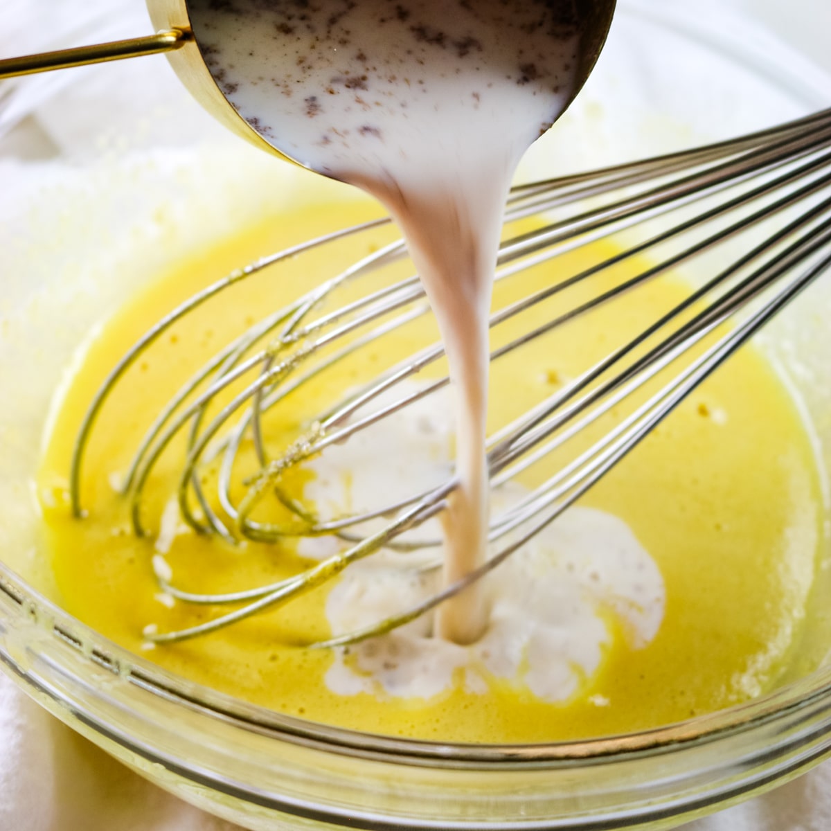 Close-up of a mixing bowl with a yellow batter being whisked. A measuring cup is pouring a stream of creamy liquid into the bowl, and several metal whisks are partially submerged in the mixture.
