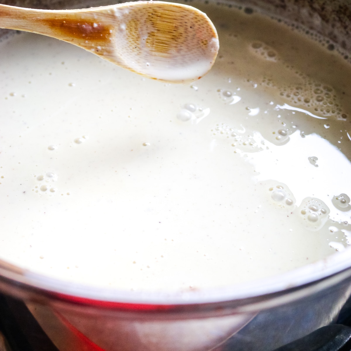 A close-up view of a creamy, light-colored mixture simmering in a pot. Small bubbles dot the surface, and a wooden spoon rests across the top, coated with the mixture.