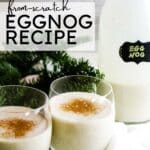 Two glasses of creamy homemade eggnog sprinkled with cinnamon, placed on a white cloth with a bottle of eggnog in the background and festive greenery around.