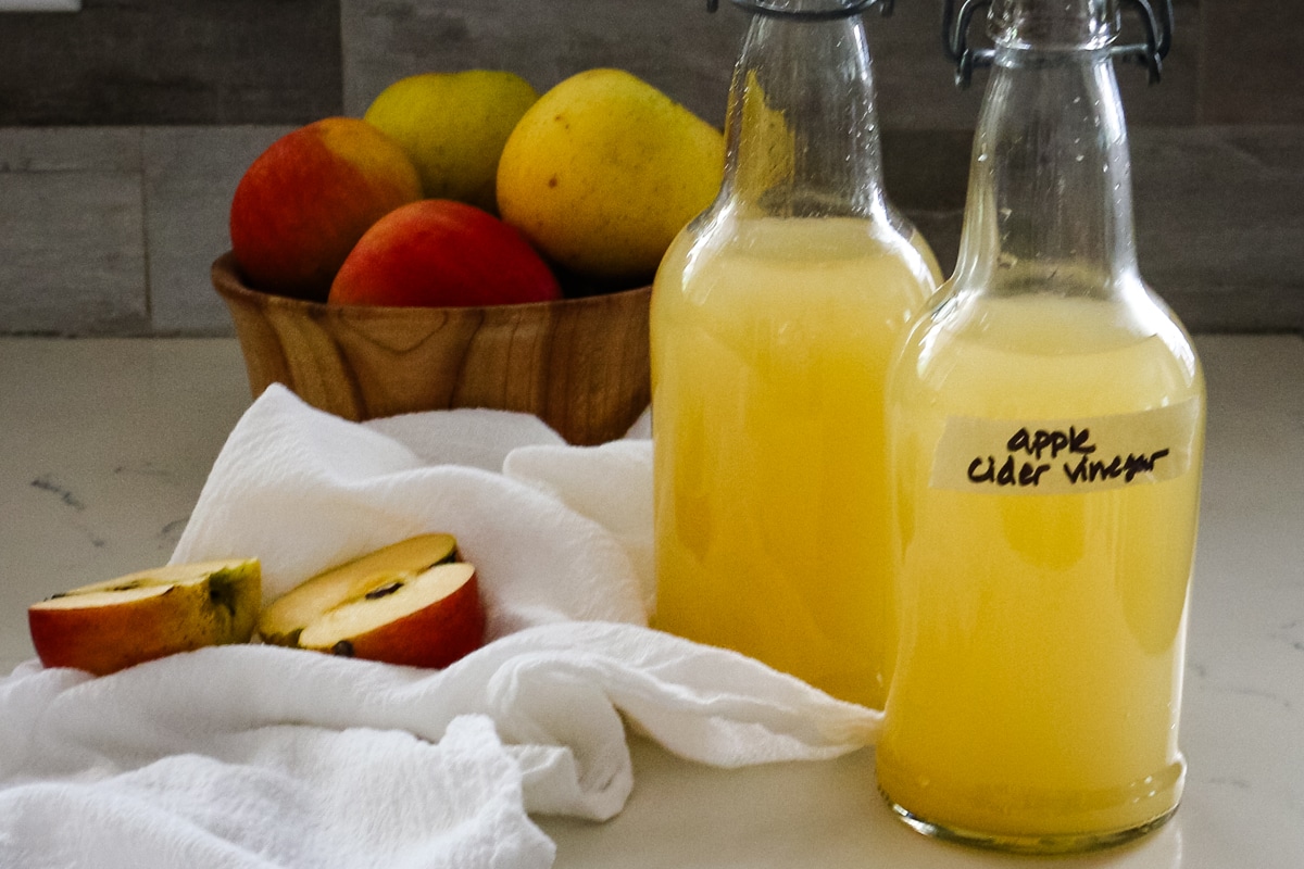 two swing-top bottles of homemade apple cider vinegar next to a bowl of fresh apples.