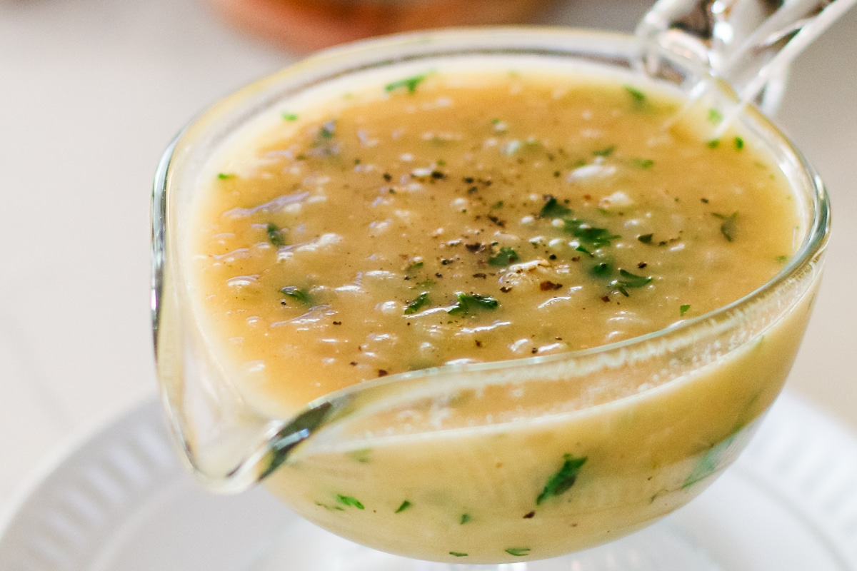 glass gravy boat filled with light brown onion gravy, topped with fresh parsley and ground black pepper, resting on a white plate.