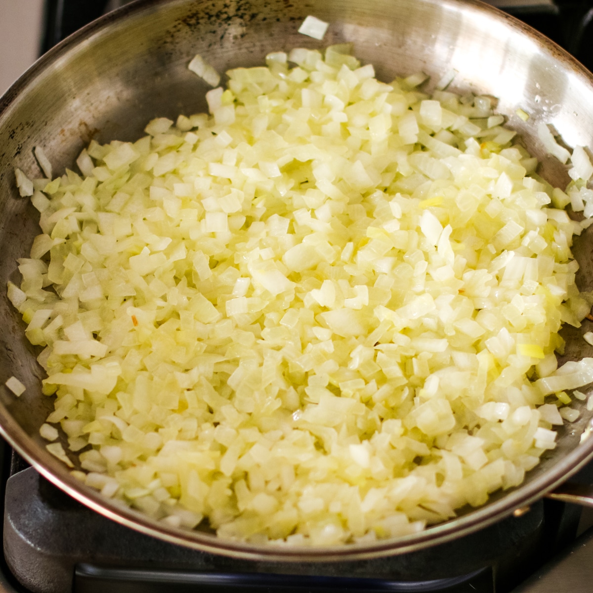 large amount of finely chopped white onions sautéing in a stainless steel skillet on a stovetop.