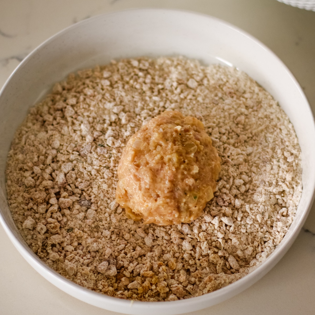 raw, round chicken mixture patty being coated in coarse breadcrumbs inside a white bowl.