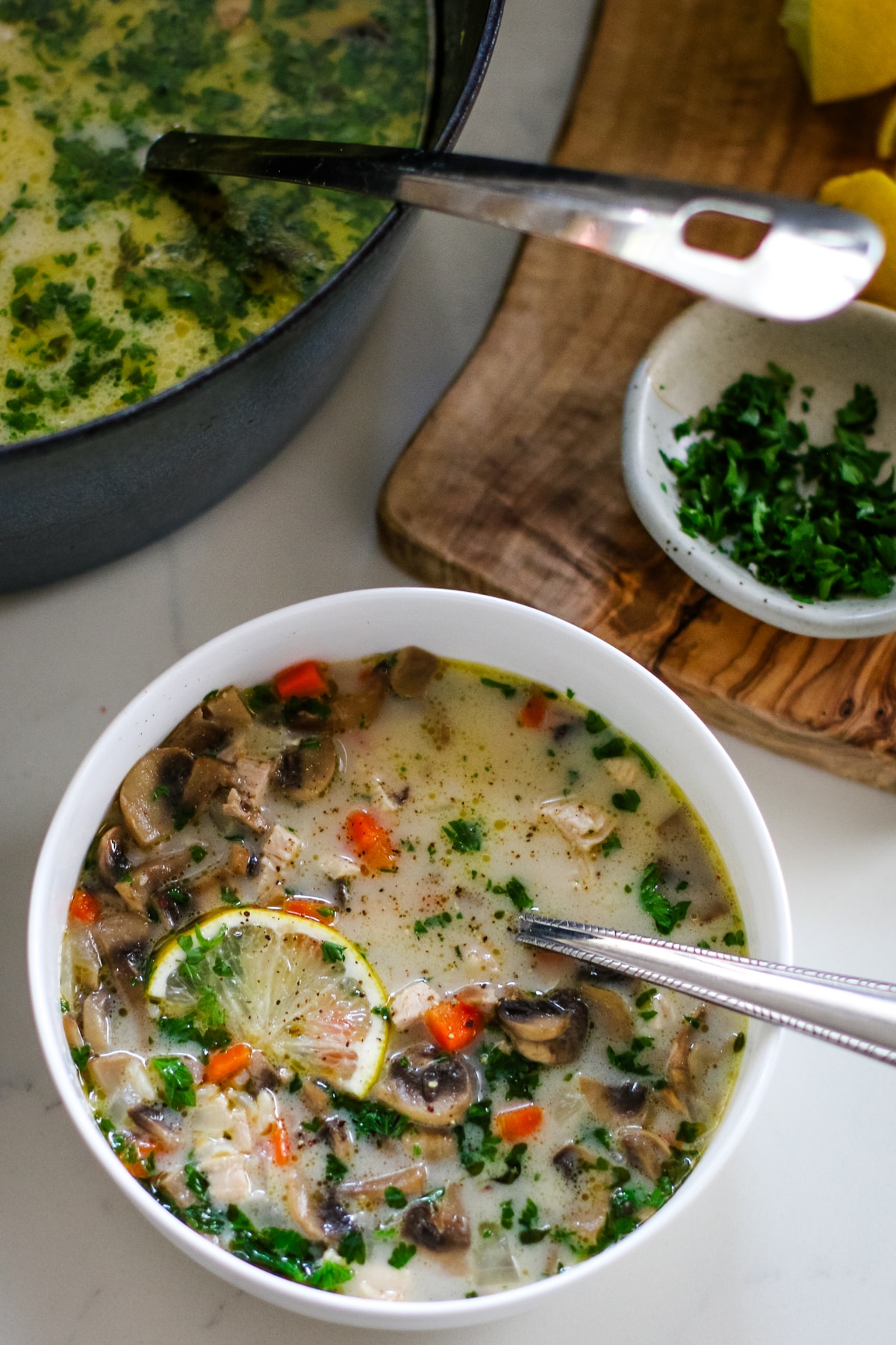 Turkey soup in a white bowl wtih a spoon and a slice of lemon, next to a cutting board and a pot full of turkey soup.