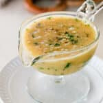 glass gravy boat filled with light brown onion gravy, topped with fresh parsley and ground black pepper, resting on a white plate.
