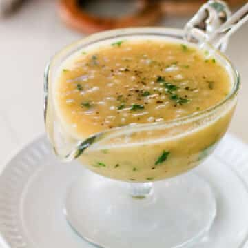 glass gravy boat filled with light brown onion gravy, topped with fresh parsley and ground black pepper, resting on a white plate.