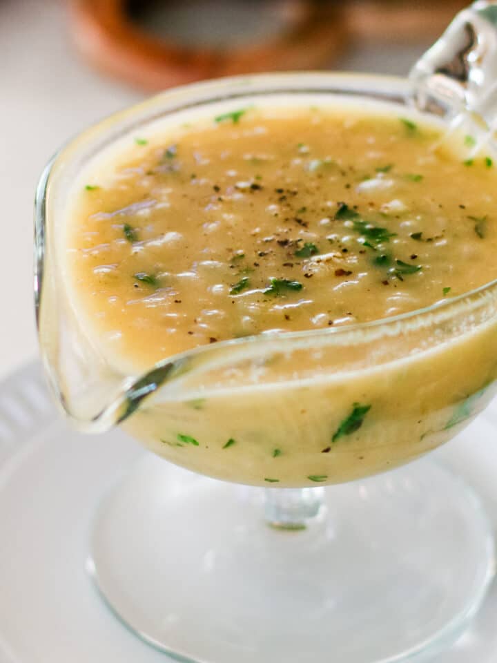 glass gravy boat filled with light brown onion gravy, topped with fresh parsley and ground black pepper, resting on a white plate.