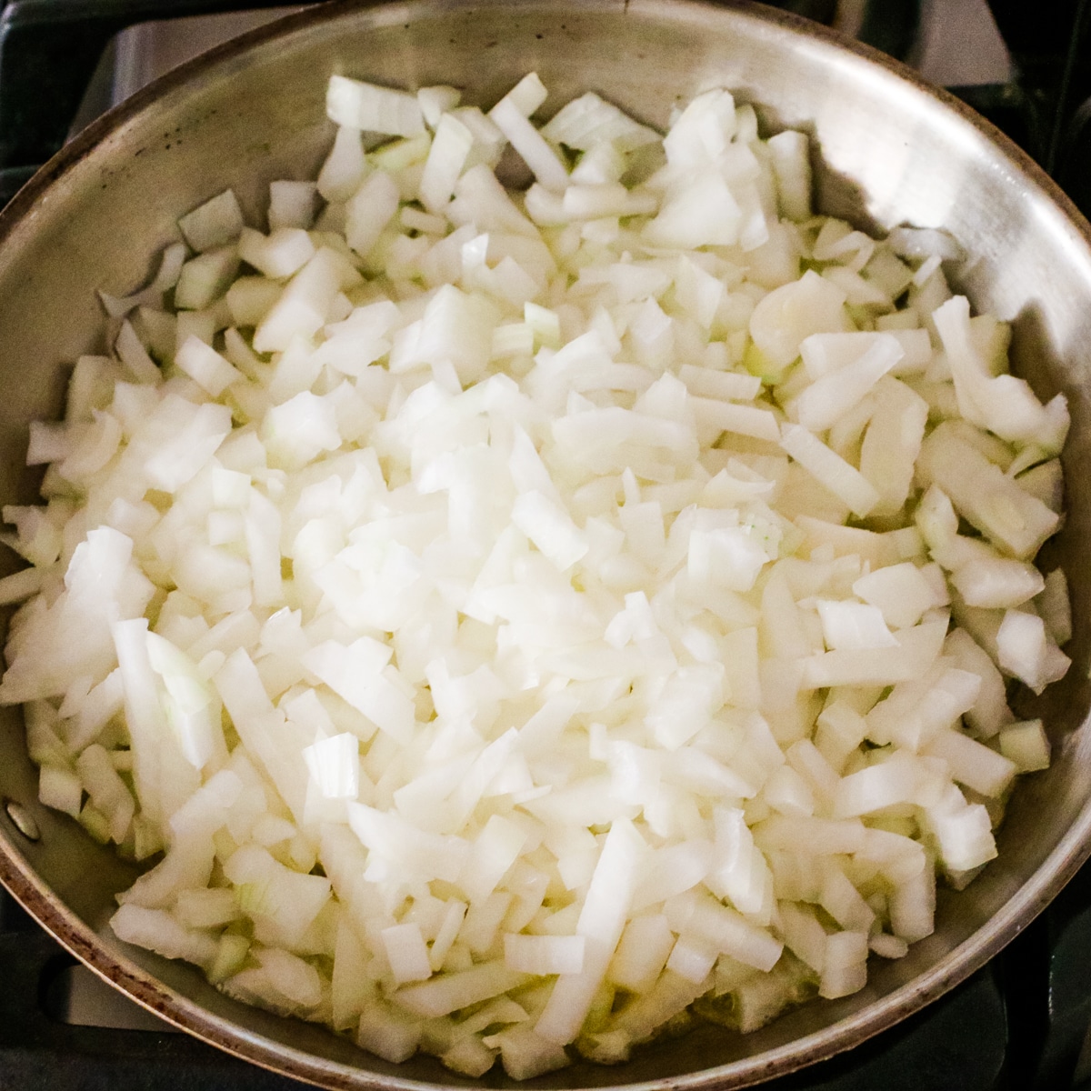 stainless steel pan on a stovetop, filled with chopped onions.