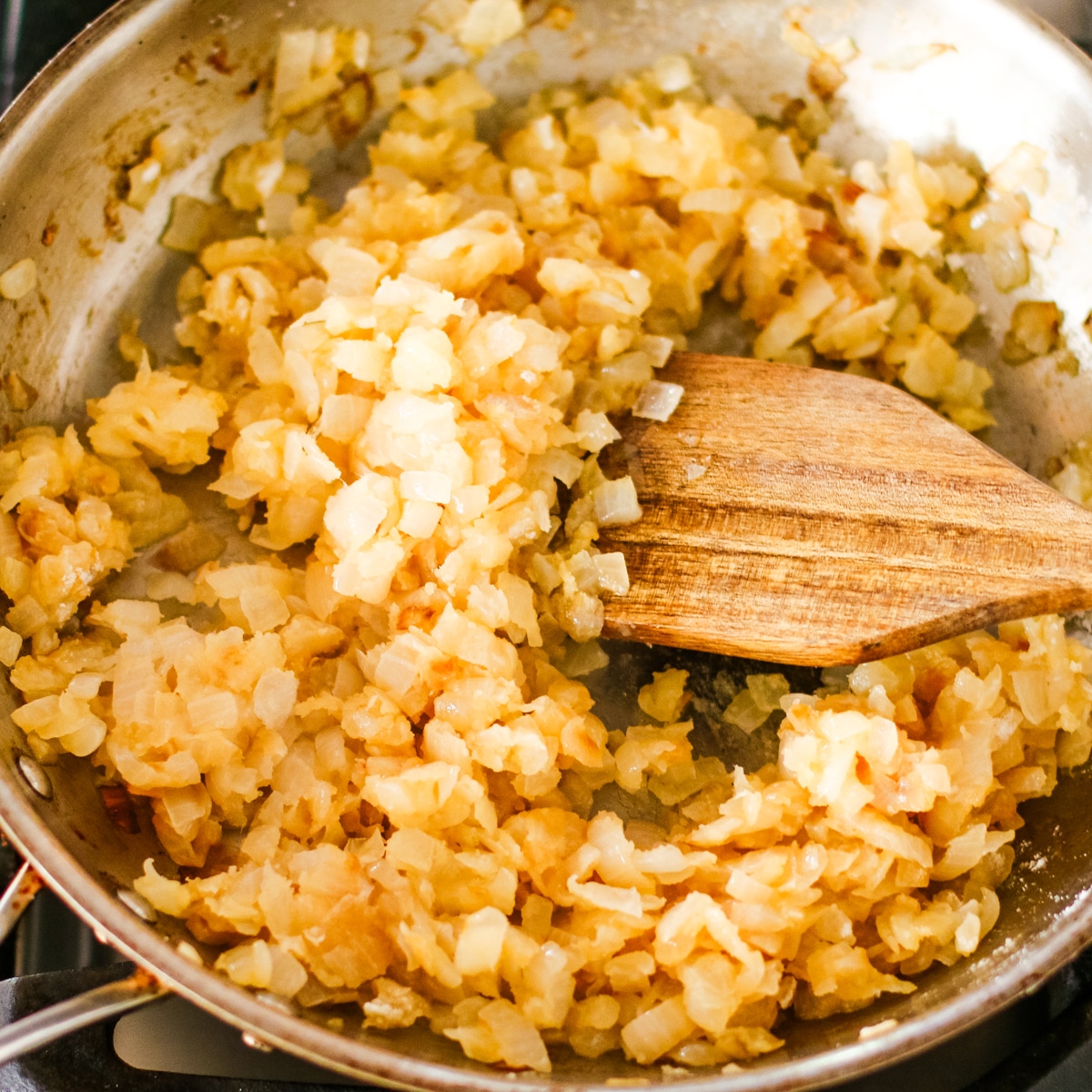 stainless steel pan on a stovetop, filled with chopped onions, being stirred with a wooden spoon.