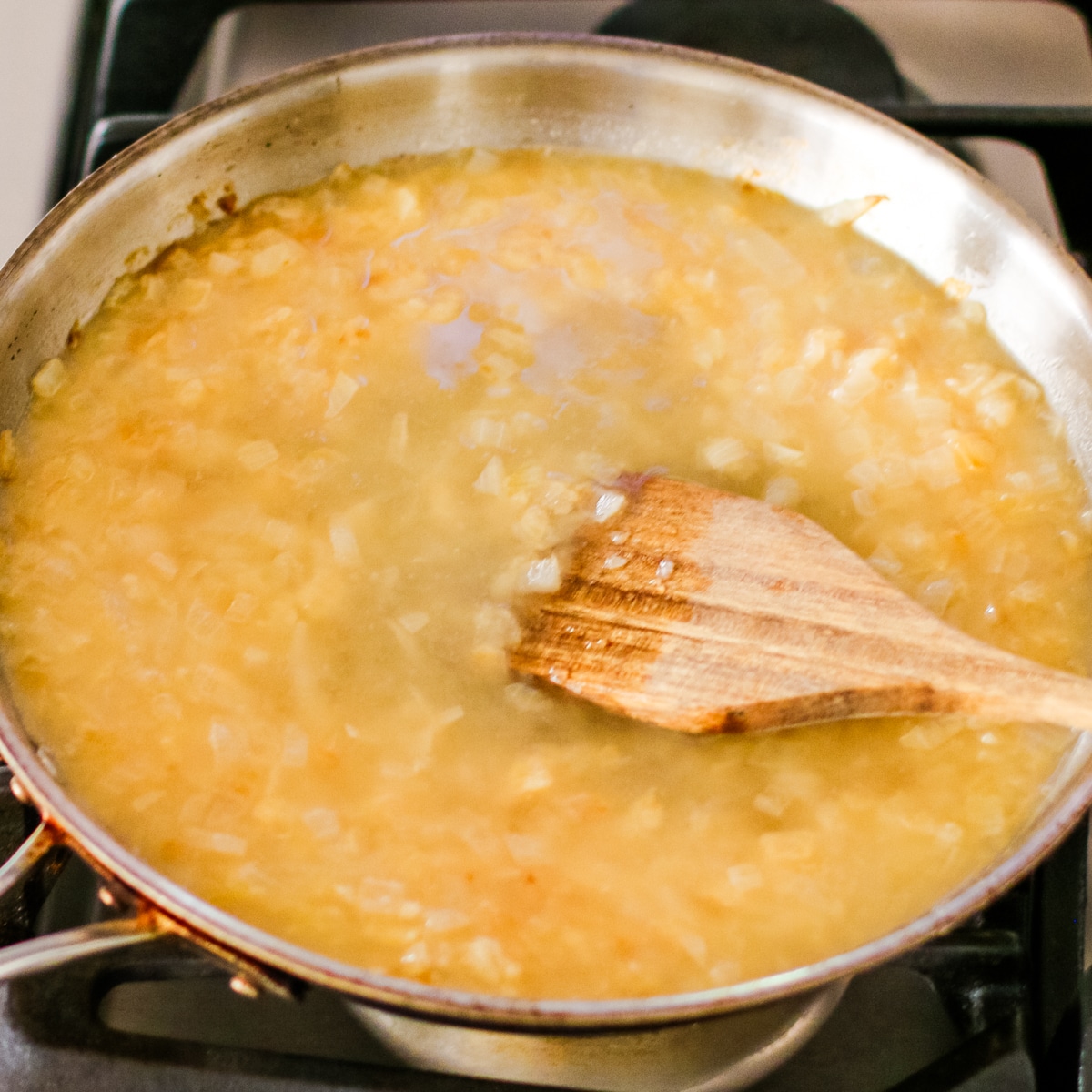 stainless steel pan on a stovetop, filled with a simmering, light brown liquid and chopped onions, being stirred with a wooden spoon.