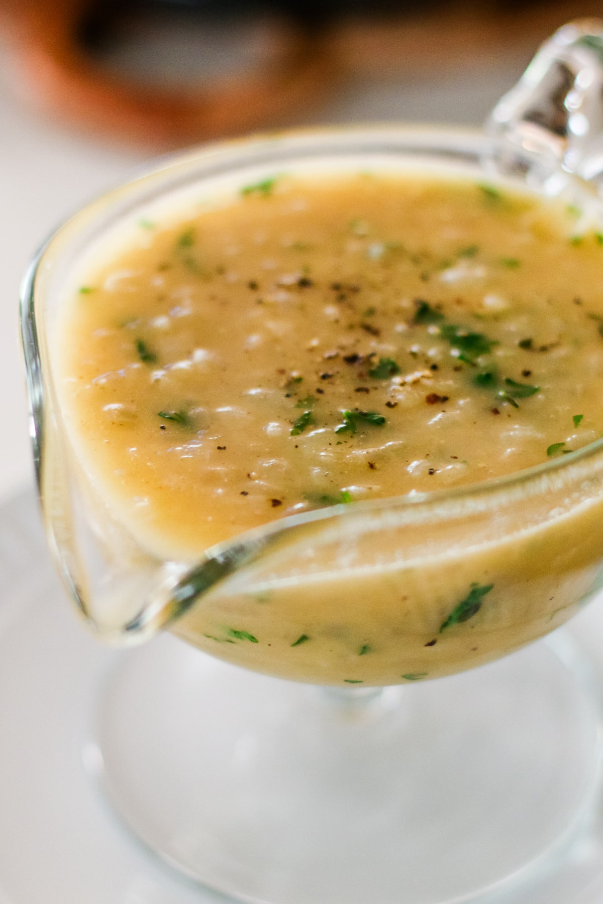 glass gravy boat filled with light brown onion gravy, topped with fresh parsley and ground black pepper, resting on a white plate.