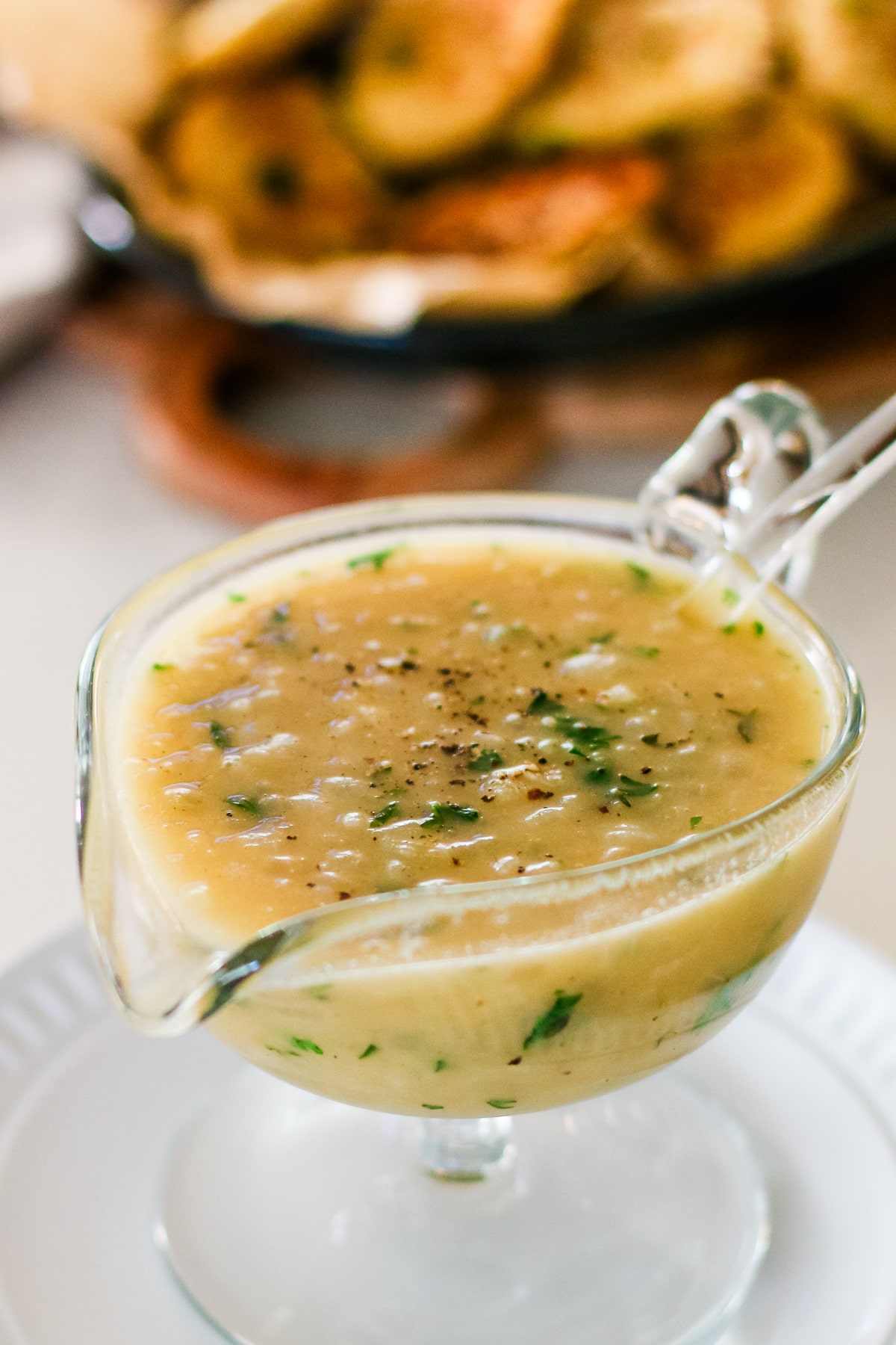 glass gravy boat filled with light brown onion gravy, topped with fresh parsley and ground black pepper, resting on a white plate.