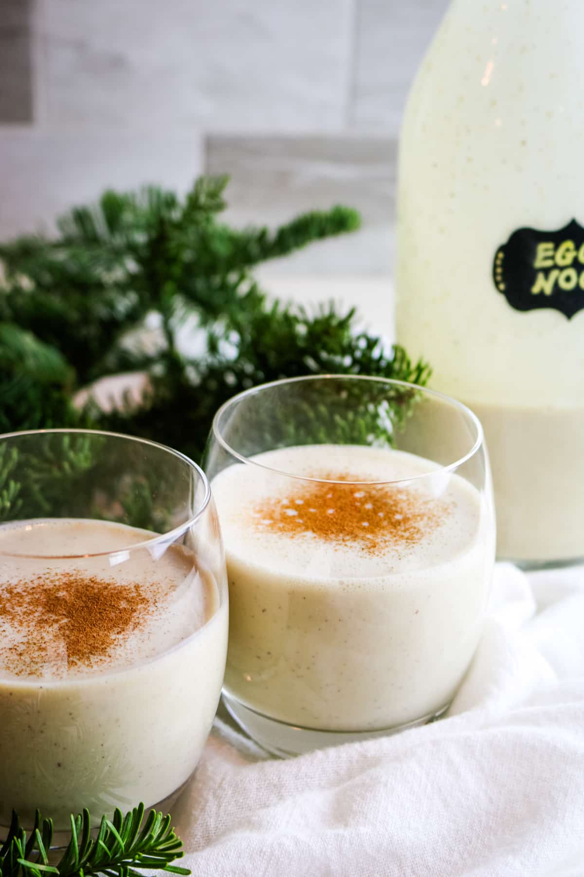 Two glasses of creamy eggnog sit on a white cloth, each topped with a sprinkle of cinnamon.