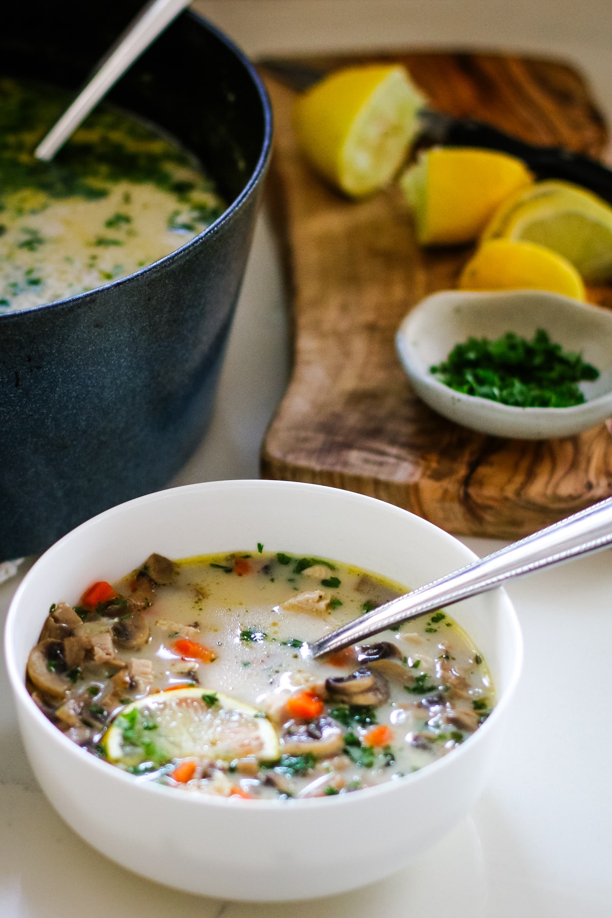 Turkey soup in a white bowl wtih a spoon and a slice of lemon, next to a cutting board and a pot full of turkey soup.