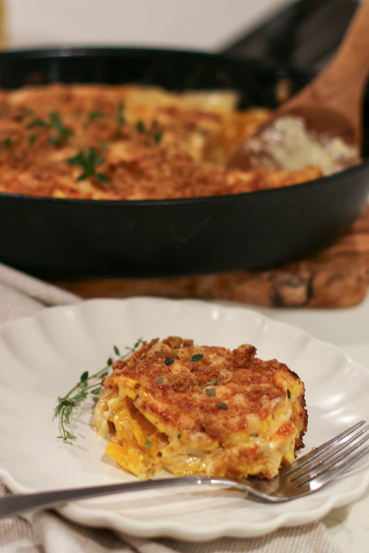 serving of baked delicata squash gratin on a plate with a fork, with the full gratin in a baking dish blurred in the background.