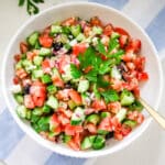 large bowl of Greek salad being tossed with a herb vinaigrette, with the dressing visible coating the cucumbers and tomatoes.