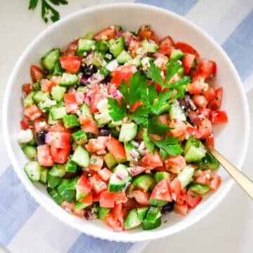 large bowl of Greek salad being tossed with a herb vinaigrette, with the dressing visible coating the cucumbers and tomatoes.