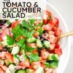 large bowl of Greek salad being tossed with a herb vinaigrette, with the dressing visible coating the cucumbers and tomatoes.