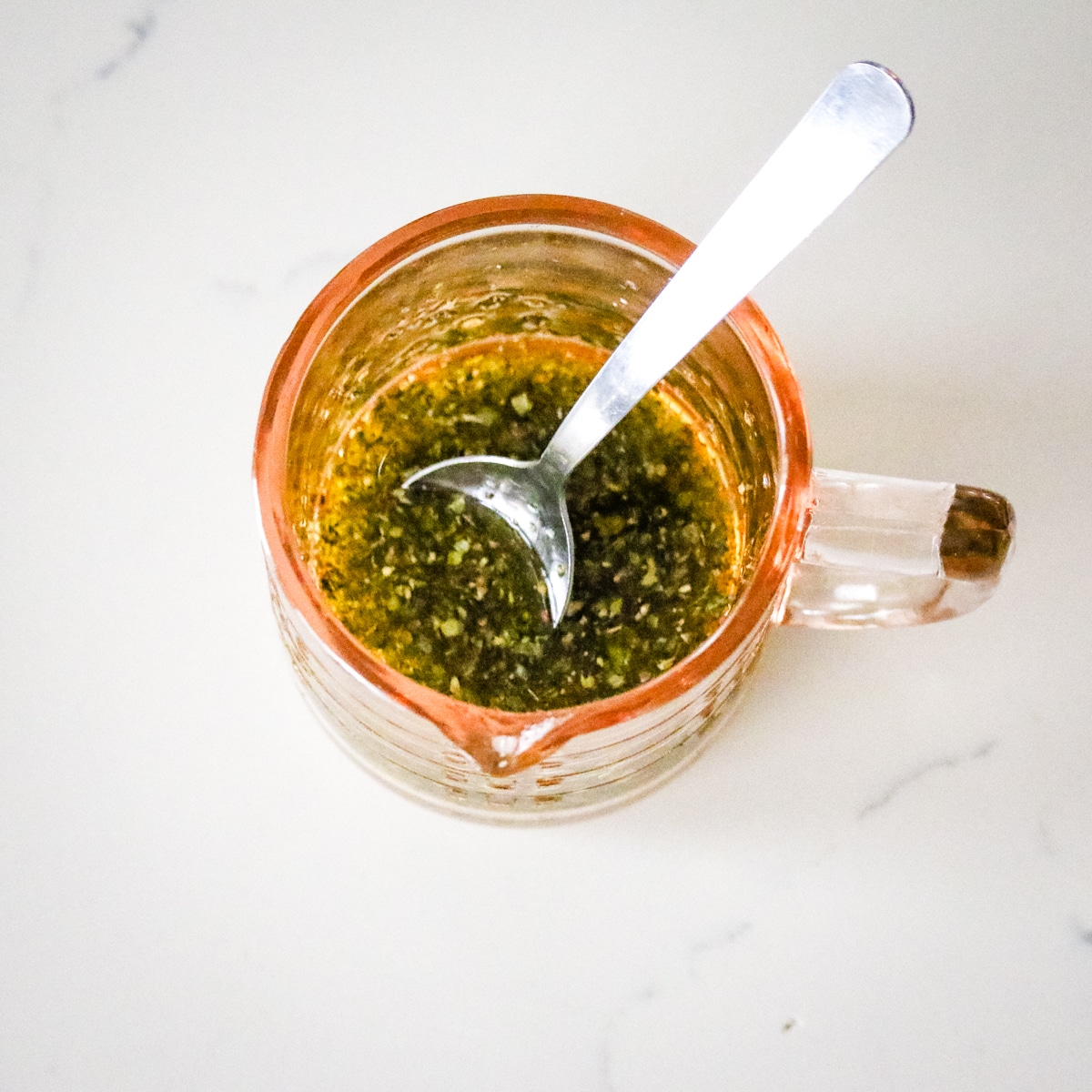 small peach-colored glass pitcher containing a dark green mixture of herbs, olive oil, and lemon juice with a silver spoon resting inside.