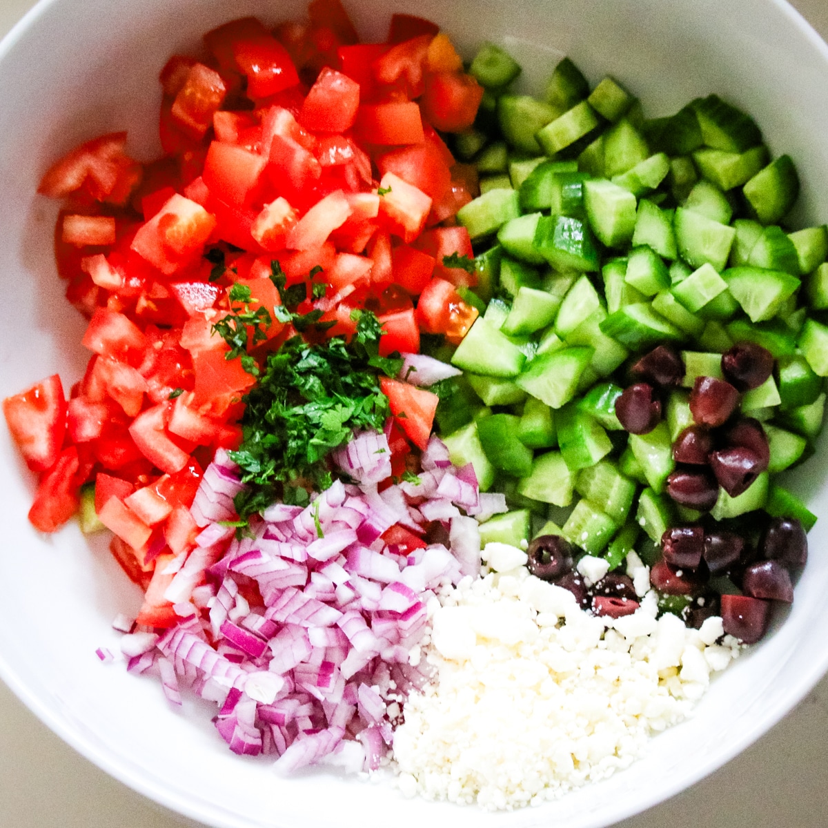 large bowl filled with diced cucumbers, tomatoes, red onions, feta cheese, and olives, ready to be tossed.