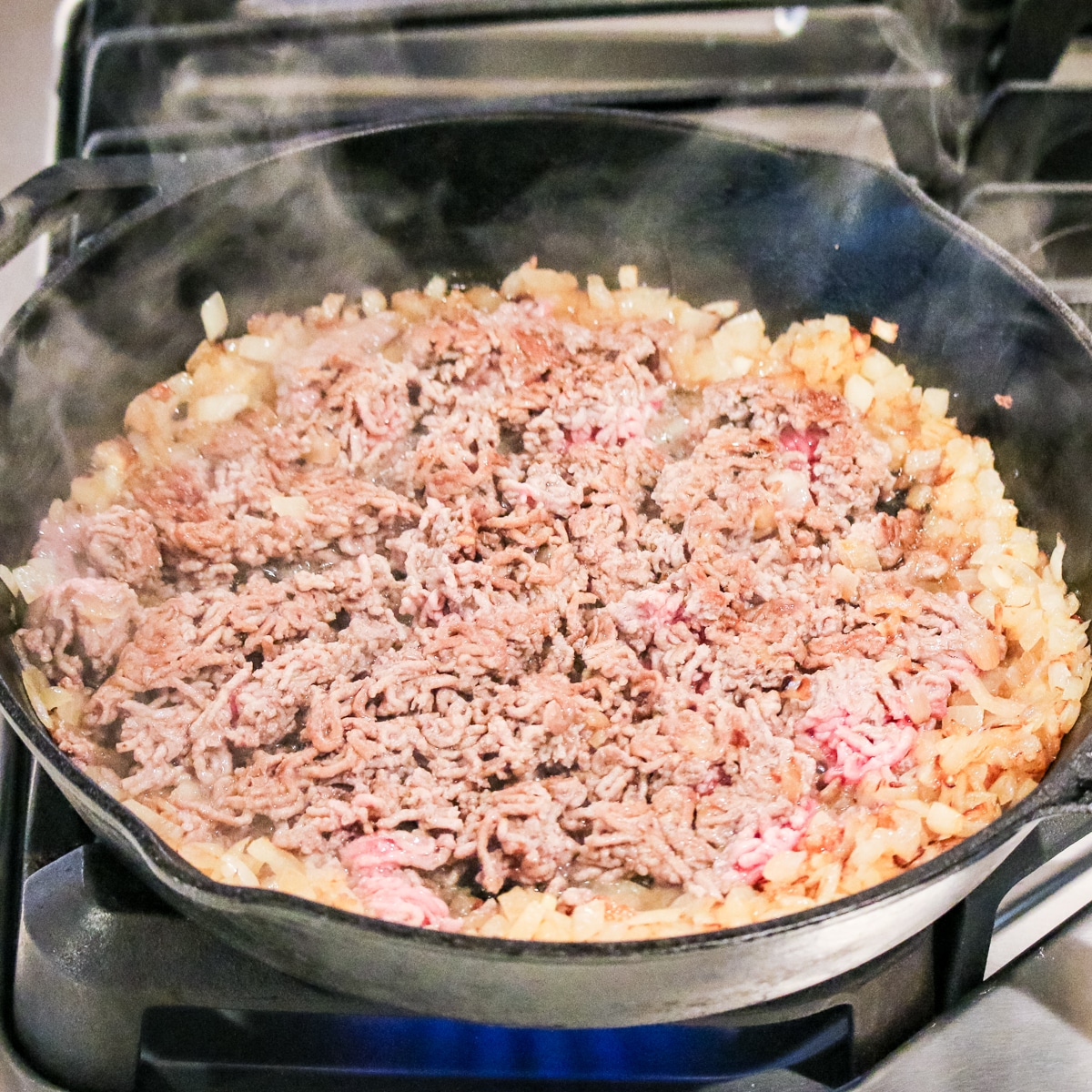 Ground beef is browning with diced onions in a black skillet.