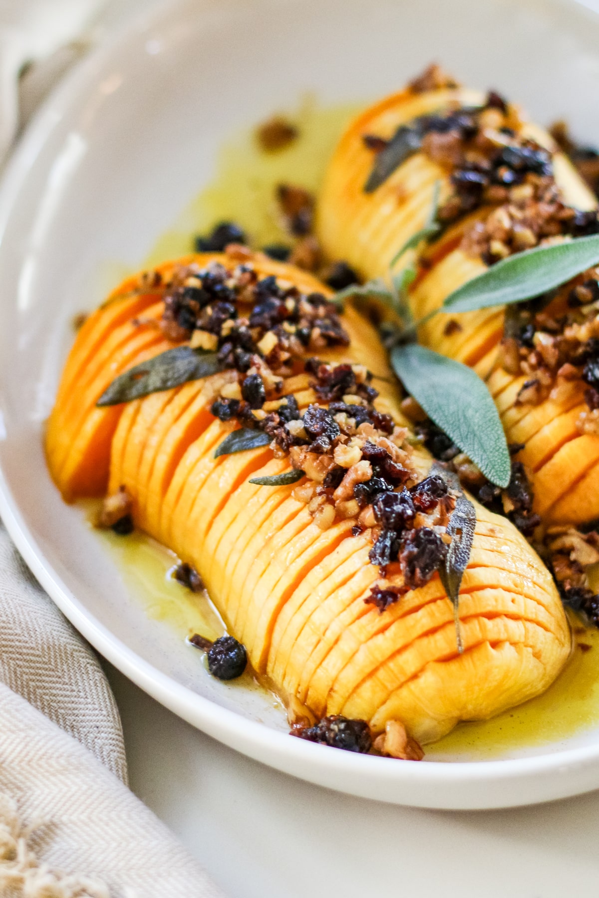 sliced hasselback butternut squash drizzled with melted butter and finished with a crunchy nut-and-dried-fruit topping, garnished with fresh sage on a white platter.