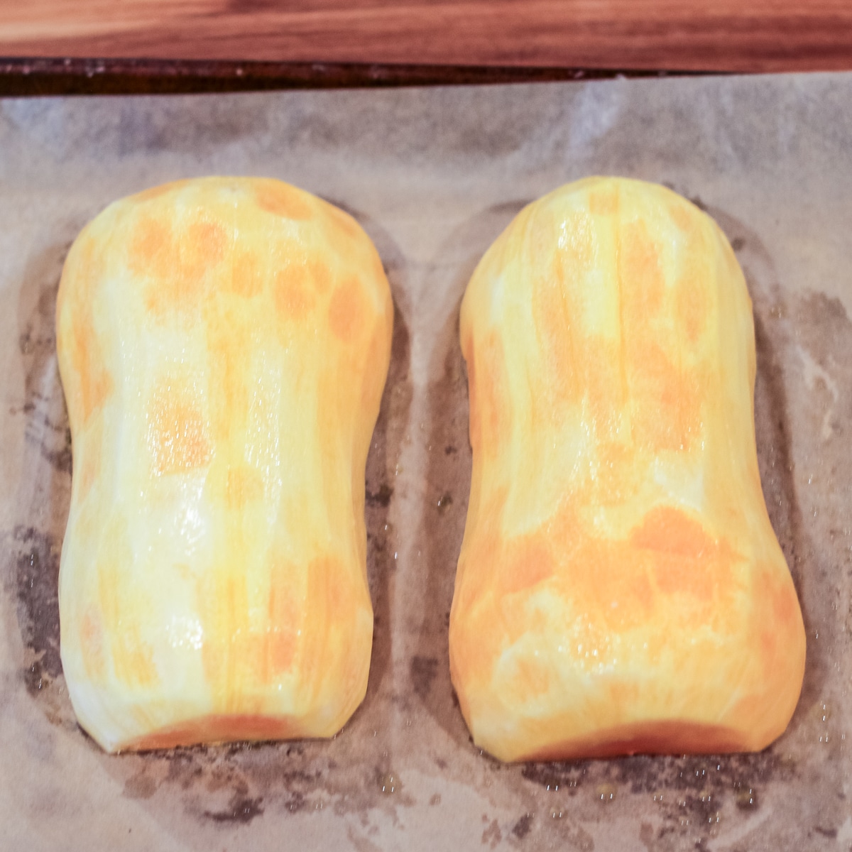 Two peeled and halved butternut squash pieces, brushed with oil, on a parchment-lined baking sheet.
