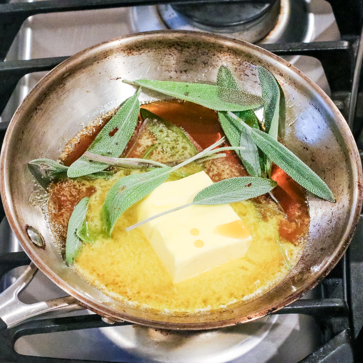 a stainless steel pan on a stovetop contains a melting stick of butter and several fresh sage leaves in a brown-butter sauce.