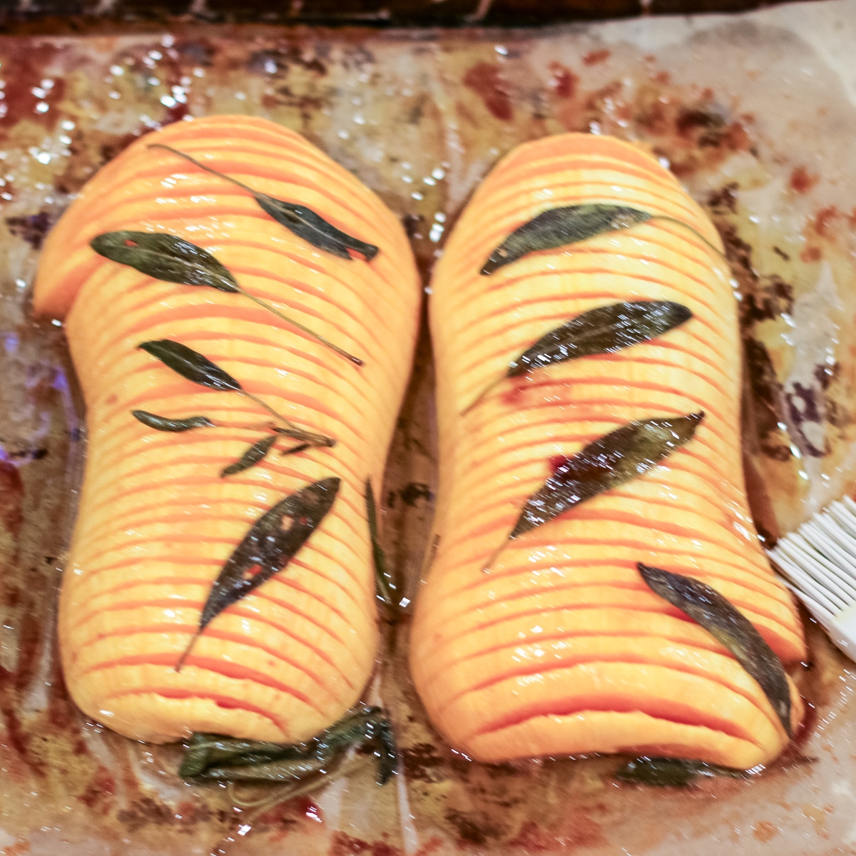 hasselback butternut squash halves topped with sage leaves, glistening with oil on a baking sheet.