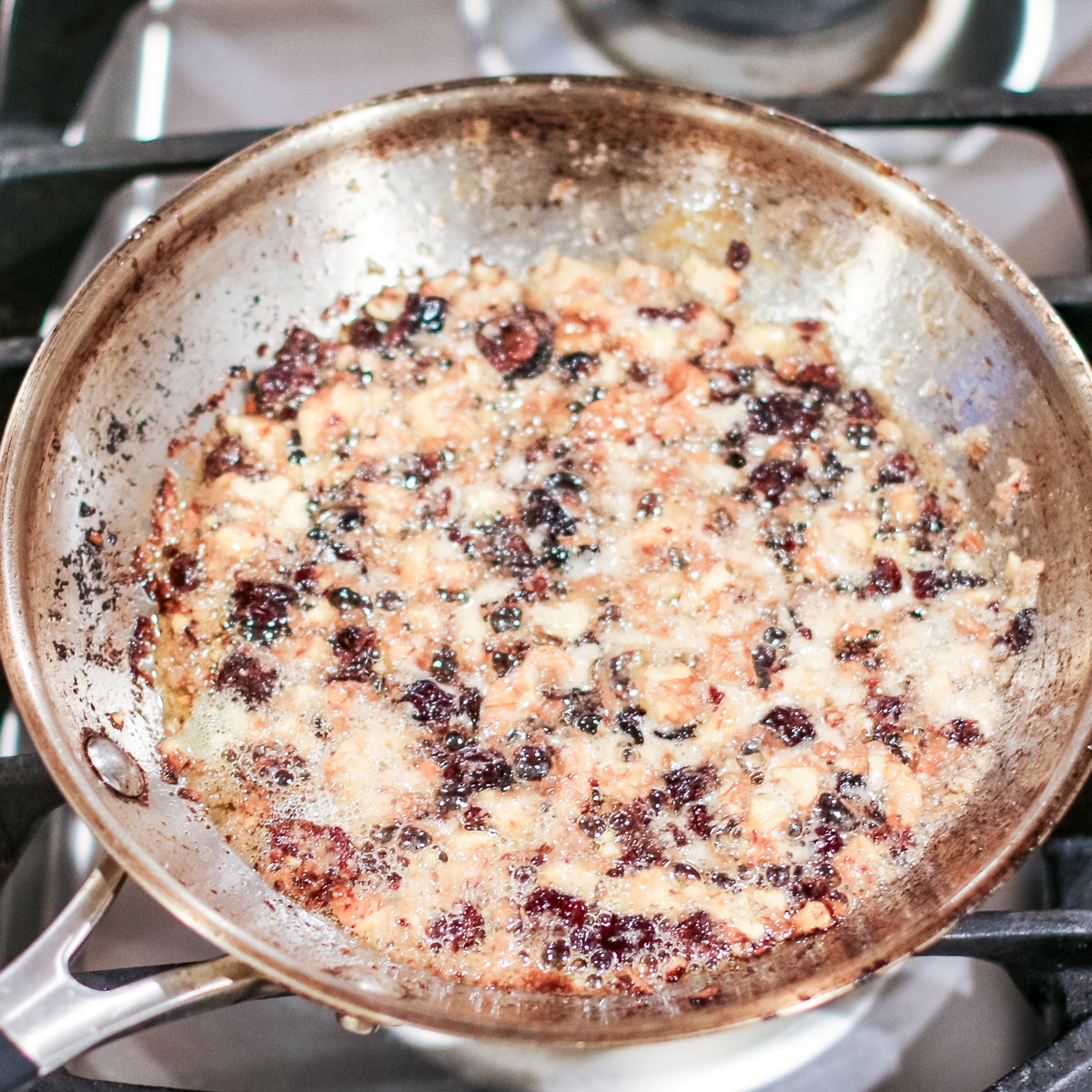 close-up of a stainless steel frying pan on a stovetop, filled with a bubbling mixture of chopped nuts and dried cranberries, likely being toasted or caramelized.