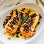 sliced hasselback butternut squash drizzled with melted butter and finished with a crunchy nut-and-dried-fruit topping, garnished with fresh sage on a white platter.