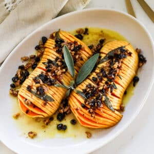 sliced hasselback butternut squash drizzled with melted butter and finished with a crunchy nut-and-dried-fruit topping, garnished with fresh sage on a white platter.
